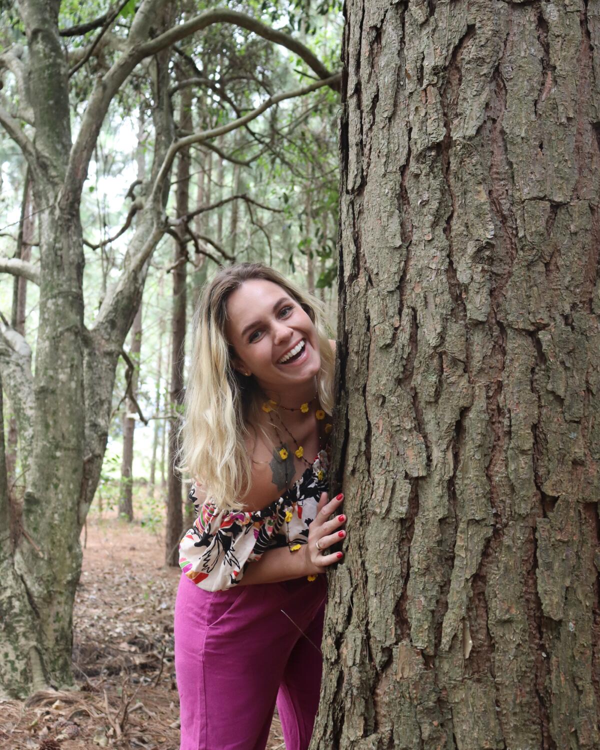 Jordana Müller smiling warmly, peeking around a tree trunk in the forest
